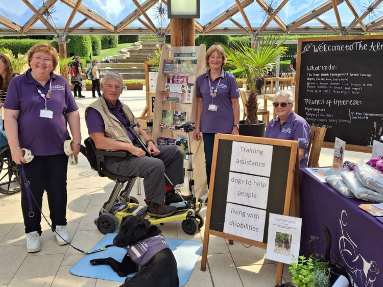 Shows 4 people and one black Labrador (canine partner) stood in front of their Canine Partners fundraising stand at a garden centre.
