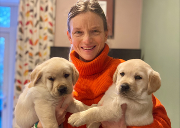 Lady in orange jumper holding two yellow lab pups one in each arm