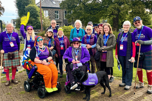A group of people are outdoors, all wearing Canine Partner merchandise, and are smiling at the camera. There is an assistance dog with his partner at the front.