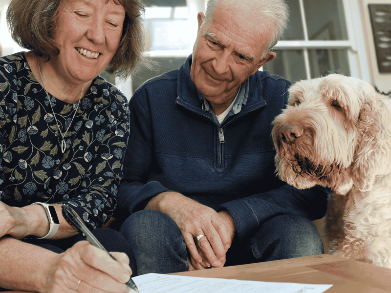 A woman signing her will, with her husband and a dog look on.